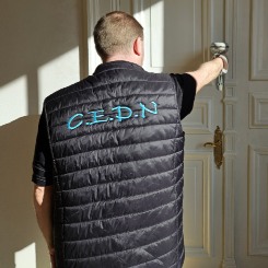 Homme portant un gilet noir C.E.D.N. ouvrant une porte blanche ornée dans un intérieur lumineux