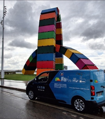 Camionnette stationnée devant une grande sculpture colorée en conteneurs empilés près de l’eau sous ciel couvert