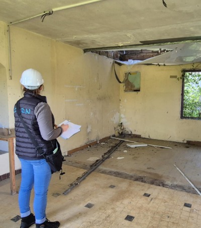 Femme avec casque de chantier inspectant une pièce dégradée aux murs abîmés et plafond endommagé